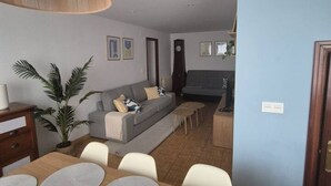 Ein Wohnzimmer mit einer Couch, einem Couchtisch, einem Esstisch und Stühlen. An der Wand hängt eine Uhr und auf dem Boden steht eine Pflanze.