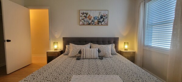 A bedroom with a patterned bedspread, two bedside lamps, a framed floral artwork above the bed, and a window with blinds.