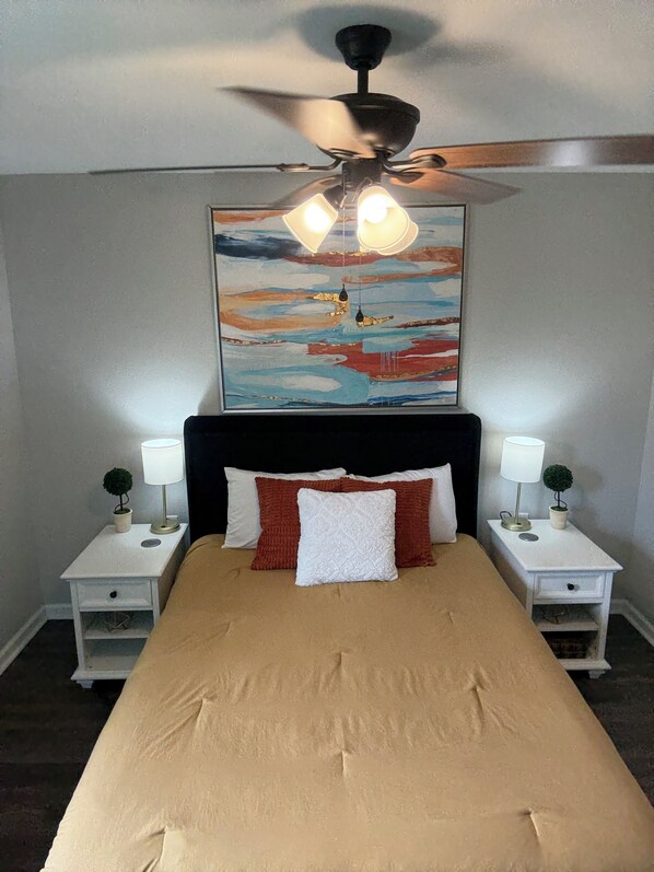 A bedroom with a bed, two bedside tables with lamps, a ceiling fan, and a painting on the wall.