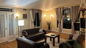 Ett vardagsrum med en soffa, ett soffbord och en fåtölj. Det finns gardiner vid fönstren, en lampa och en tavla på väggen.