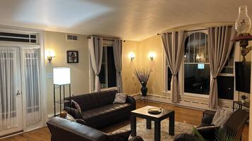 Un salon avec un canapé, une table basse et un fauteuil. Il y a des rideaux aux fenêtres, une lampe et un tableau au mur.