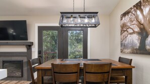 Un coin repas avec une grande table en bois, quatre chaises, une cheminée, un téléviseur et un grand tableau représentant un arbre accroché au mur.