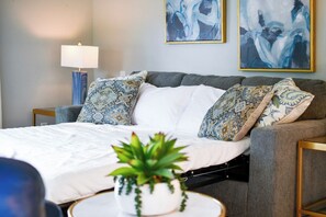 Un dormitorio moderno con un sofá cama gris, almohadas estampadas, una colcha blanca y una pequeña planta en maceta.