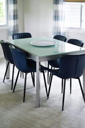 Una mesa de comedor con cubierta de vidrio y seis sillas, ubicada en una habitación con piso alfombrado y cortinas.
