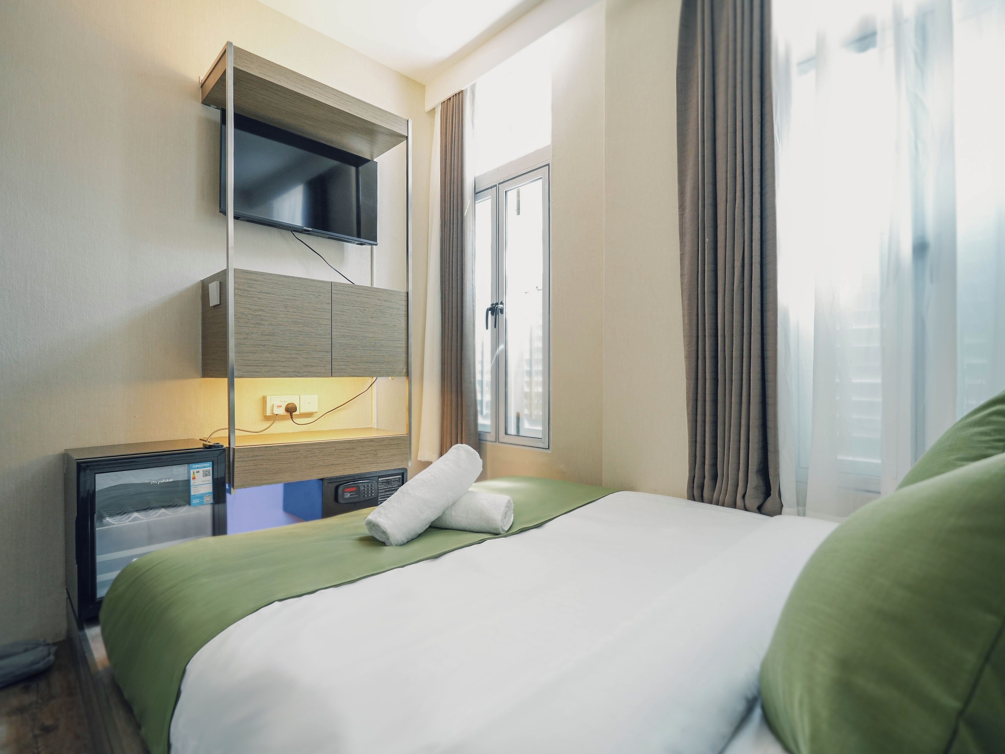 A hotel room with a bed, a TV mounted on the wall, a control panel, and a window with curtains.