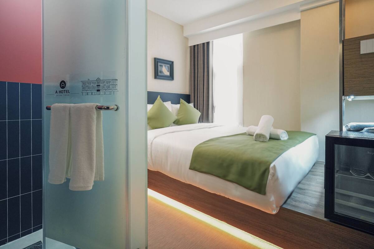 Hotelkamer met een bed, een groene plaid, een handdoek op het bed, een nachtkastje met een telefoon, en een raam met gordijnen.