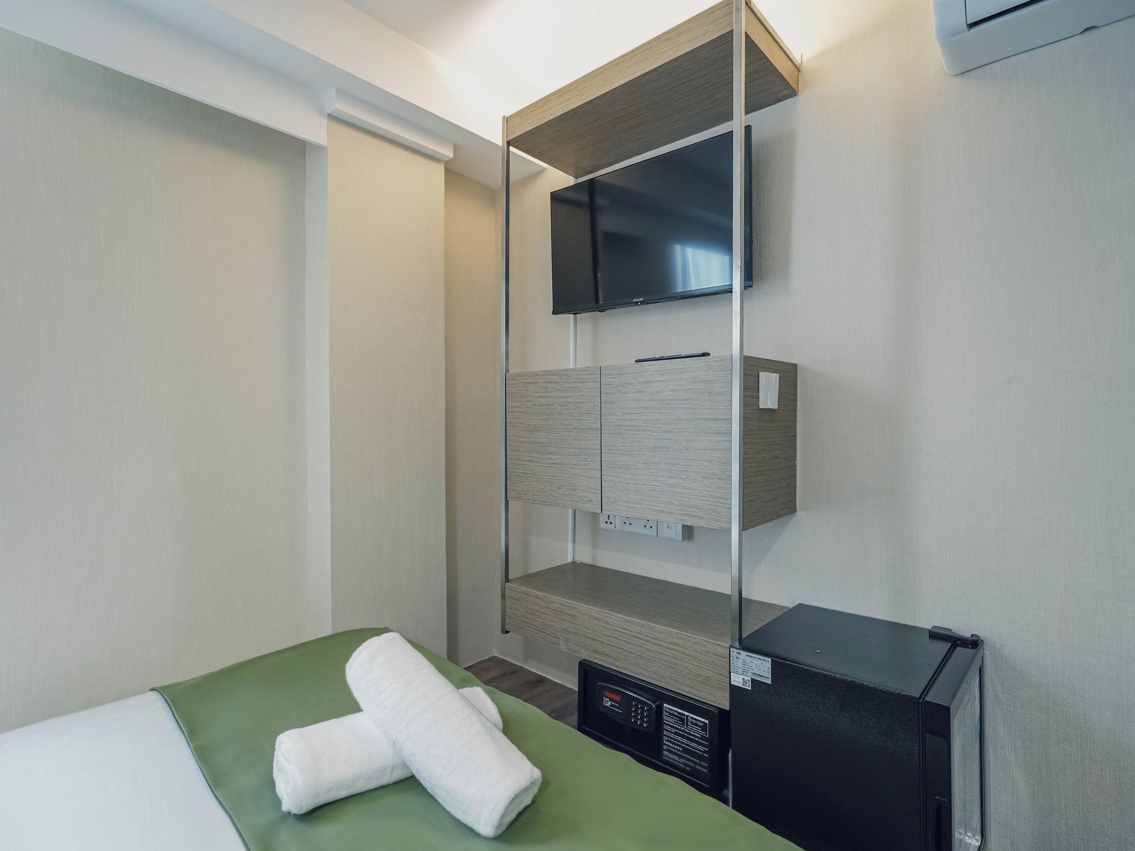 Une chambre d’hôtel avec un lit, une couvre-lit verte, deux serviettes blanches, un appareil électronique noir, une étagère en bois et un téléviseur à écran plat.