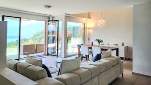 A modern living room with a sofa, dining table, and chairs. There is a sliding glass door leading to a balcony with a view of the mountains. - Residenza Solaria Endless View by Interhome (Gordola)