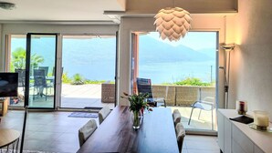 A modern dining area with a wooden table, chairs, and a vase of flowers. Large sliding glass doors lead to an outdoor patio with a view of a lake and mountains. - Residenza Solaria Endless View by Interhome (Gordola)