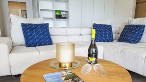 A modern living room with a round coffee table, a bottle of champagne, and two glasses. A magazine is on the table. The room has a white sofa with blue patterned pillows and a bookshelf in the background. - Residenza Solaria Endless View by Interhome (Gordola)