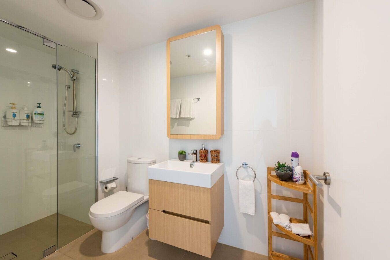 Un baño moderno con cabina de ducha de vidrio, inodoro, lavamanos con tocador de madera y espejo sobre el lavamanos.