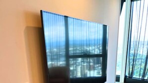 A modern living room with a large flat-screen TV mounted on a wooden console, a black speaker, and a view of a cityscape through large windows.