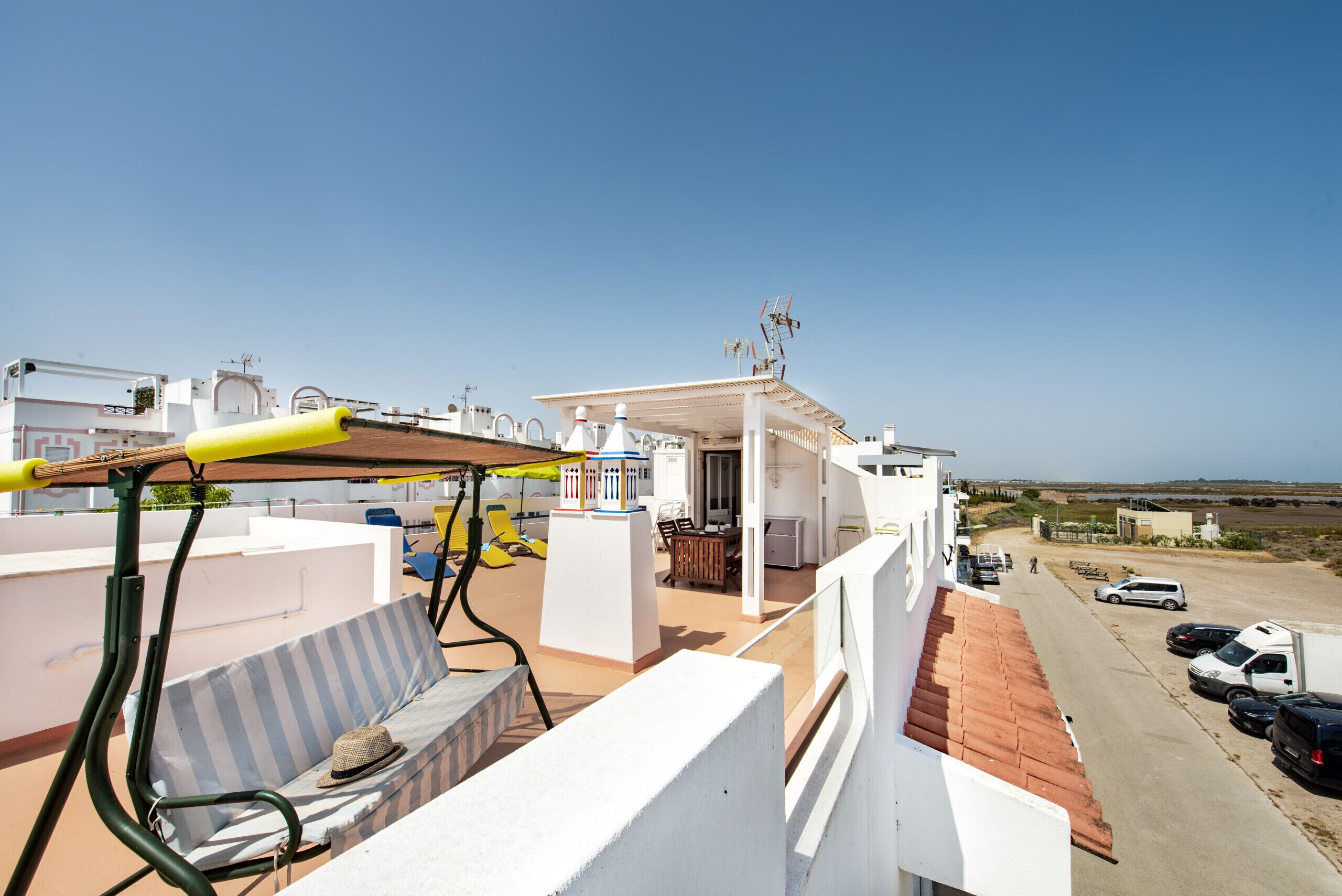 Ein Balkon mit einer gestreiften Couch, einem Tisch und einem gelben Sonnenschirm. Im Hintergrund ist ein Parkplatz und ein Gebäude zu sehen.