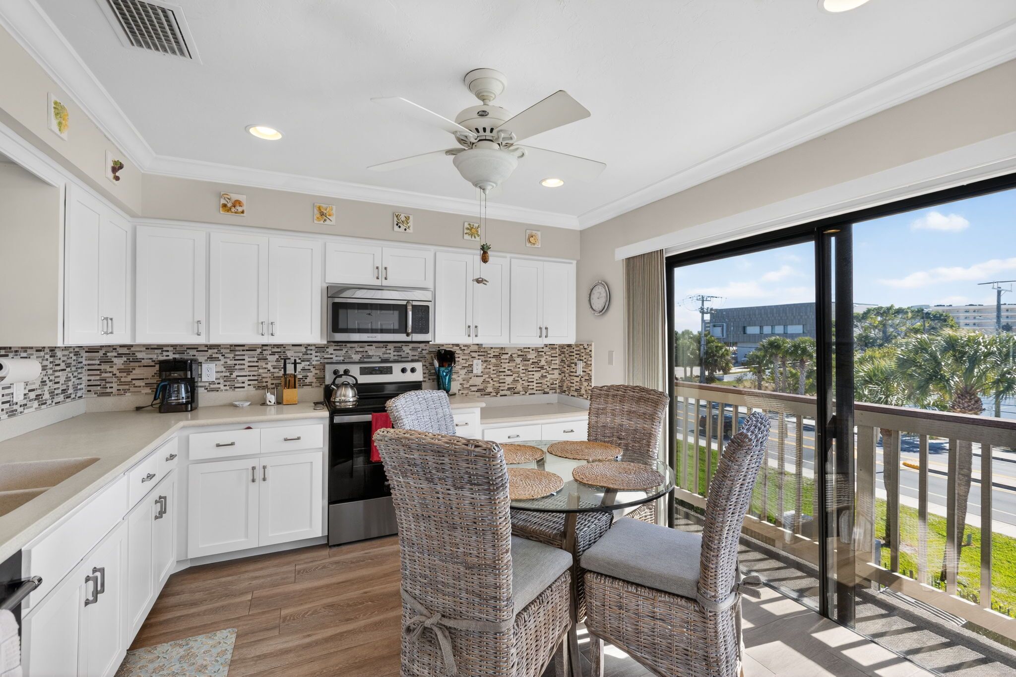 A modern kitchen with white cabinets, a microwave, and a stove. There is a dining area with wicker chairs and a glass table. A ceiling fan is visible, and the room has a large window with a view of palm trees.