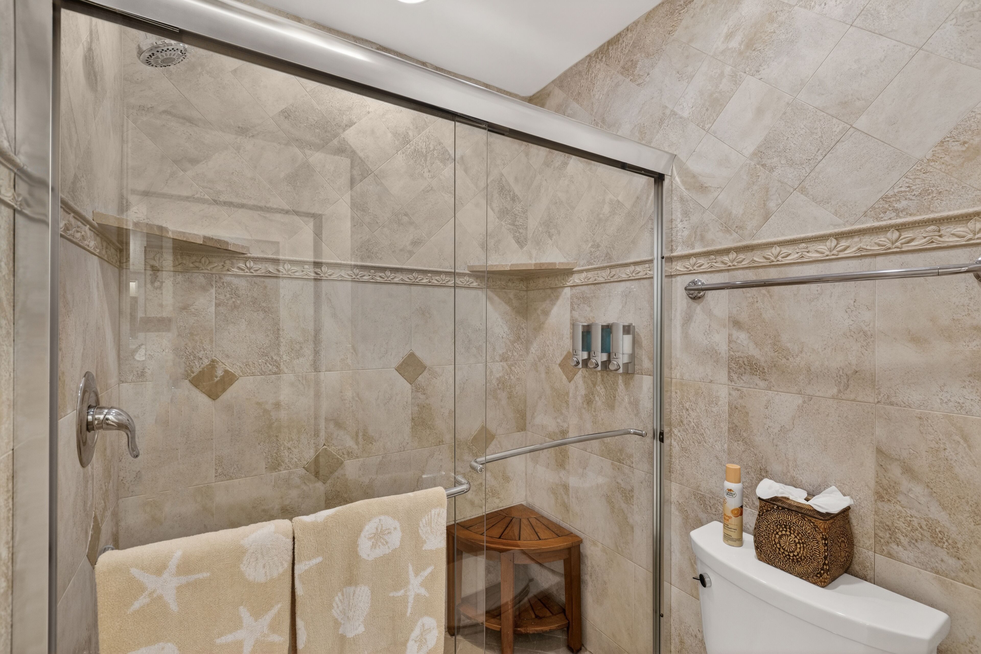 A glass-enclosed shower with a glass door, a wooden stool, a toilet with a decorative box, and a towel rack with towels.
