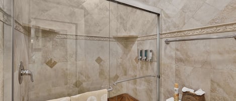 A glass-enclosed shower with a glass door, a wooden stool, a toilet with a decorative box, and a towel rack with towels.