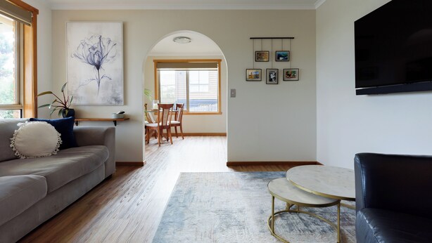 Una sala de estar con un sofá, una mesa de centro y un televisor. Se observa un comedor con sillas y una mesa al fondo.