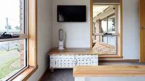 Una habitación con un ventanal grande, un televisor de pantalla plana en la pared, un banco de madera y un pequeño armario blanco con cajones.