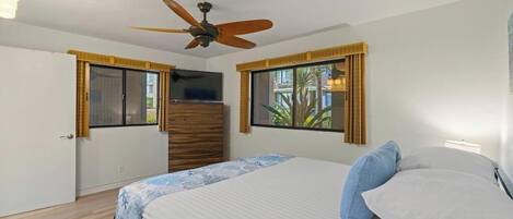 Un dormitorio con una cama, ventilador de techo, televisor y dos ventanas con cortinas.