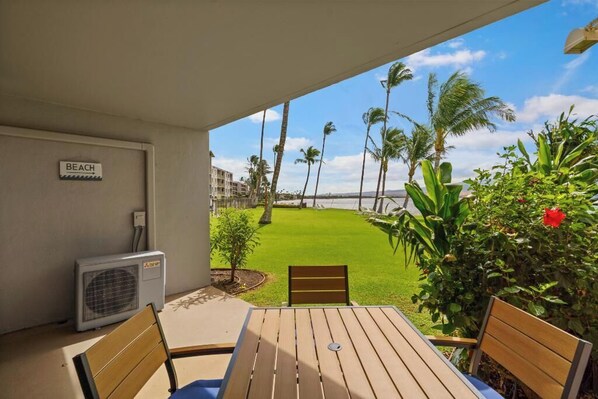 A patio with a table and chairs, a sign indicating 'BEACH', and a view of a well-maintained lawn with palm trees. - Ma'alaea Kai #102 – Peaceful Ground-Floor Oceanfront Escape with AC and Relaxing Views (Wailuku, Maui)