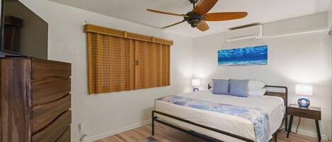 Un dormitorio con cama, ventilador de techo, ventana con persianas, aire acondicionado instalado en la pared y una mesita de noche con lámpara.