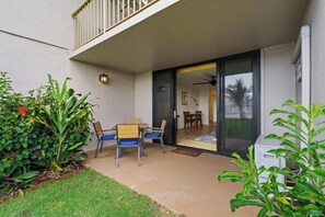 Un patio exterior con una mesa y sillas, un portillo corredizo que da a una habitación, y un pequeño jardín con plantas.
