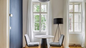 Une pièce avec une table ronde, deux chaises grises, un lampadaire et de grandes fenêtres ornées de rideaux blancs.