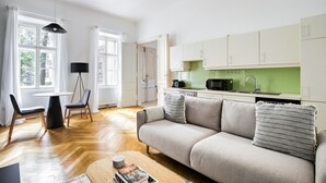 Un salon moderne comprenant un canapé, une table basse et un coin repas avec des chaises et une table. La cuisine est dotée de meubles blancs et d’un plan de travail vert.