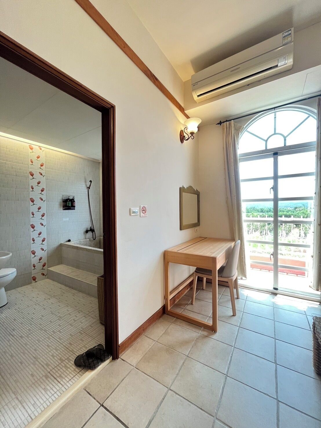 A bathroom with a toilet, shower, and a mirror. There is a wooden table and a chair in the room.