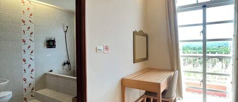 A bathroom with a toilet, shower, and a mirror. There is a wooden table and a chair in the room.