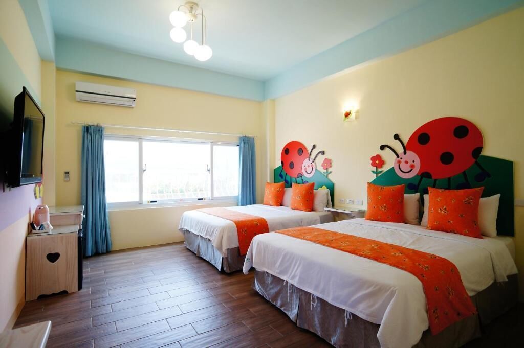 A hotel room with two beds, a TV, a window with curtains, and a wall decoration of ladybugs.