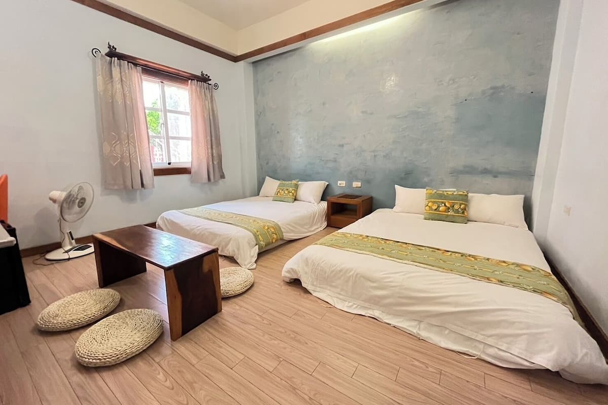 A hotel room with two beds, a small table, a fan, and a window with curtains.