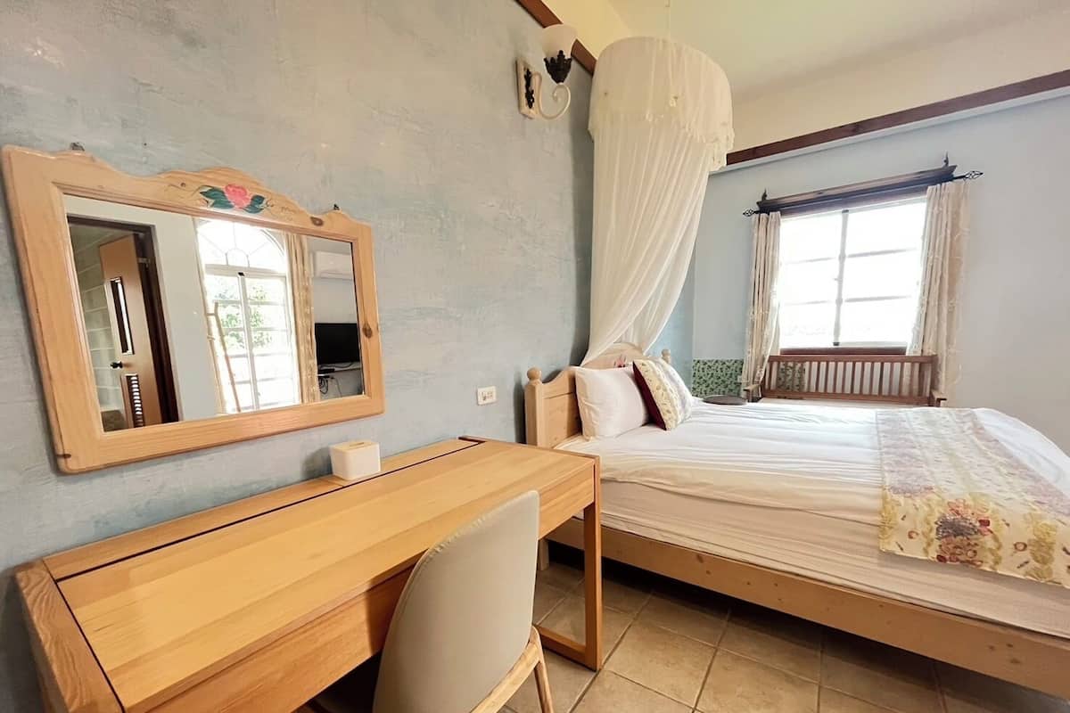 A bedroom with a wooden desk, a chair, a bed with a canopy, a window with curtains, and a wall-mounted mirror.