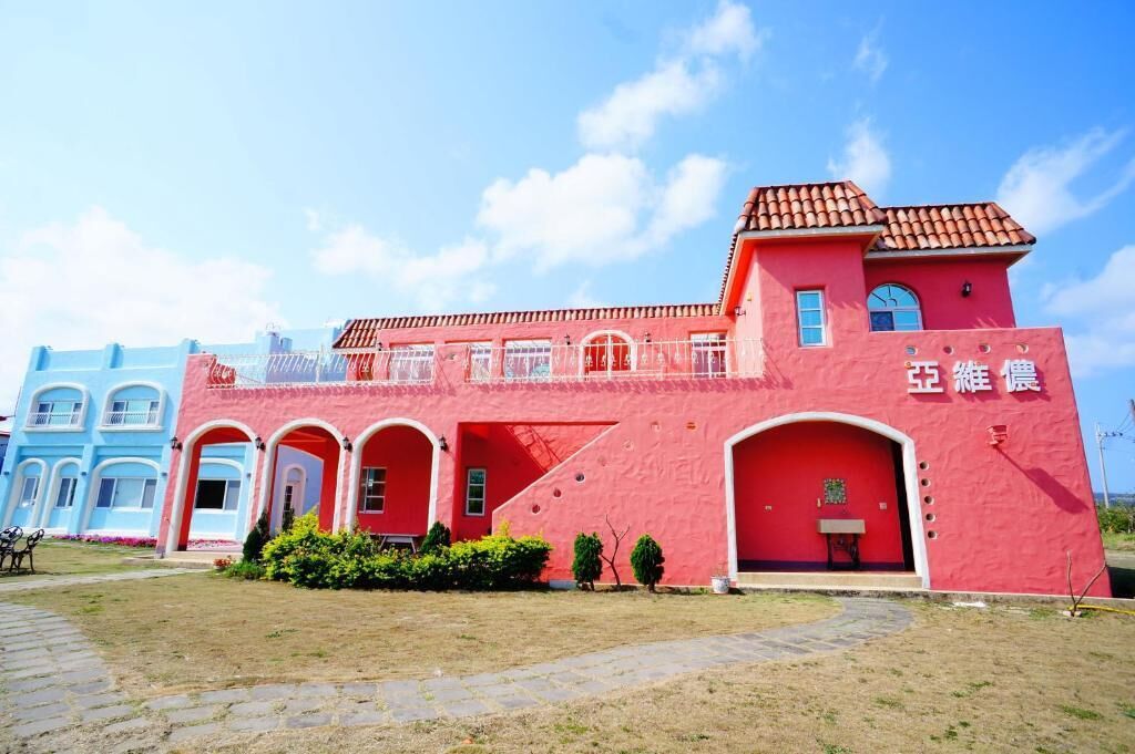 A pink building with a red door and windows, surrounded by a paved area and greenery.