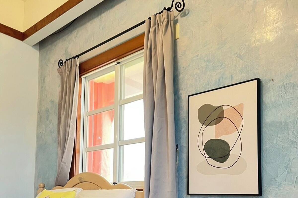 A bedroom with a bed, a bedside table, a window with curtains, and a wall-mounted abstract art piece.
