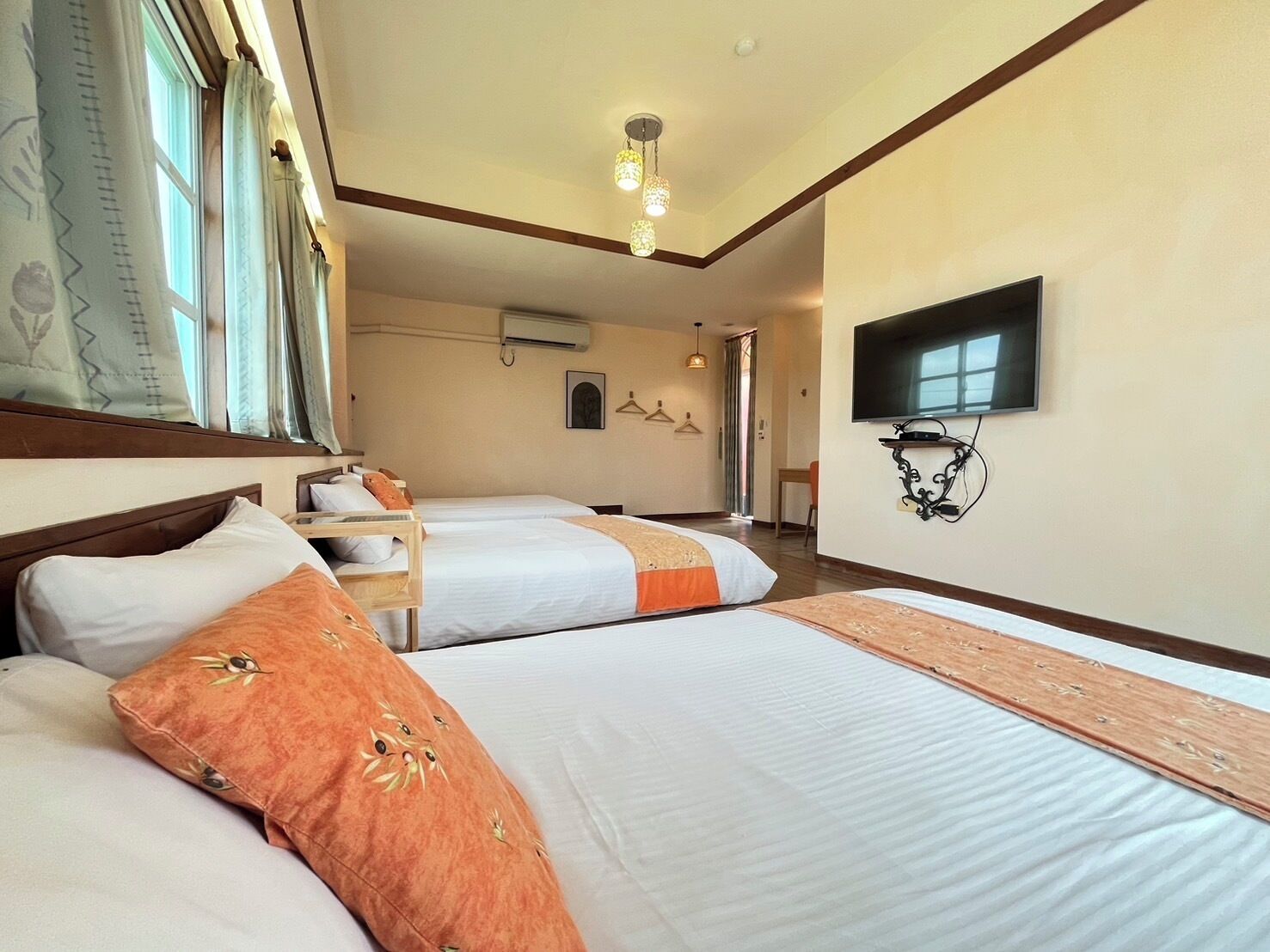 A hotel room with two beds, a TV, a hanging light fixture, and a window with curtains.