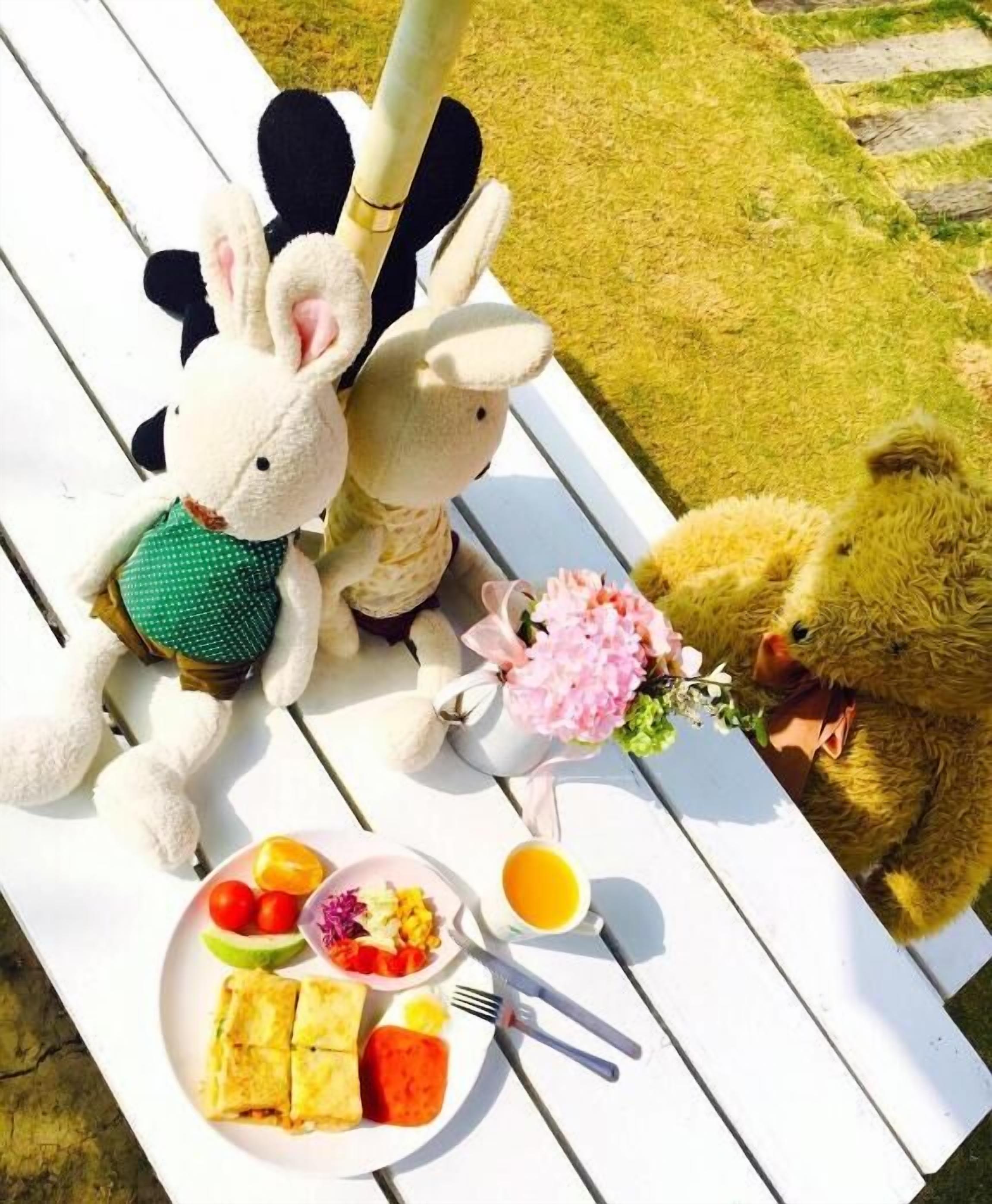 Un montaje para picnic con peluches, un plato de comida, un juego de té y un florero sobre una mesa blanca al aire libre.