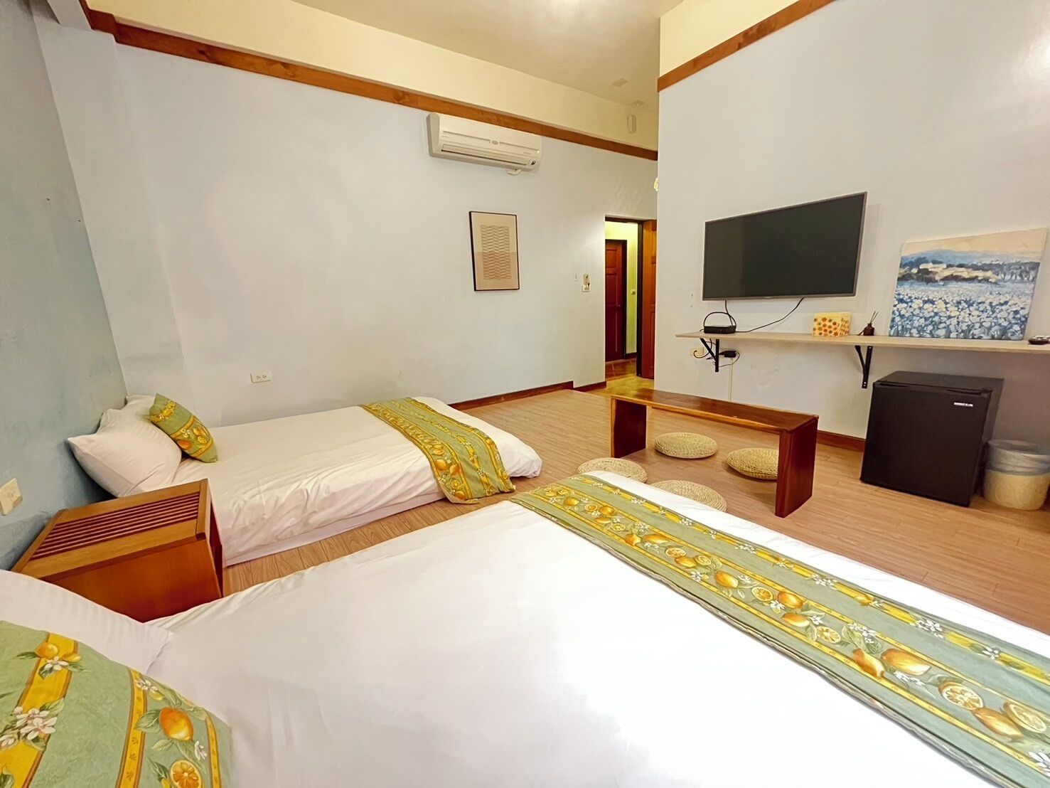 Habitación de hotel con dos camas, televisión, refrigerador pequeño y un banco de madera.