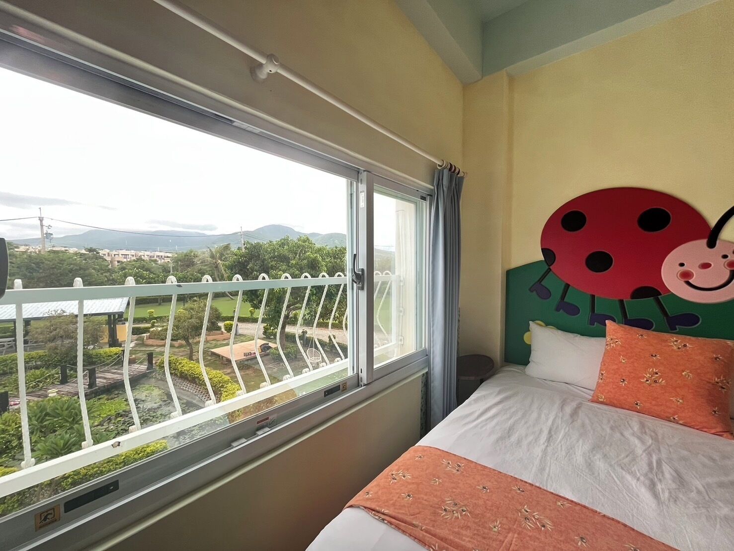 A bedroom with a large window, a bed with orange and white bedding, and a wall decoration of a ladybug.