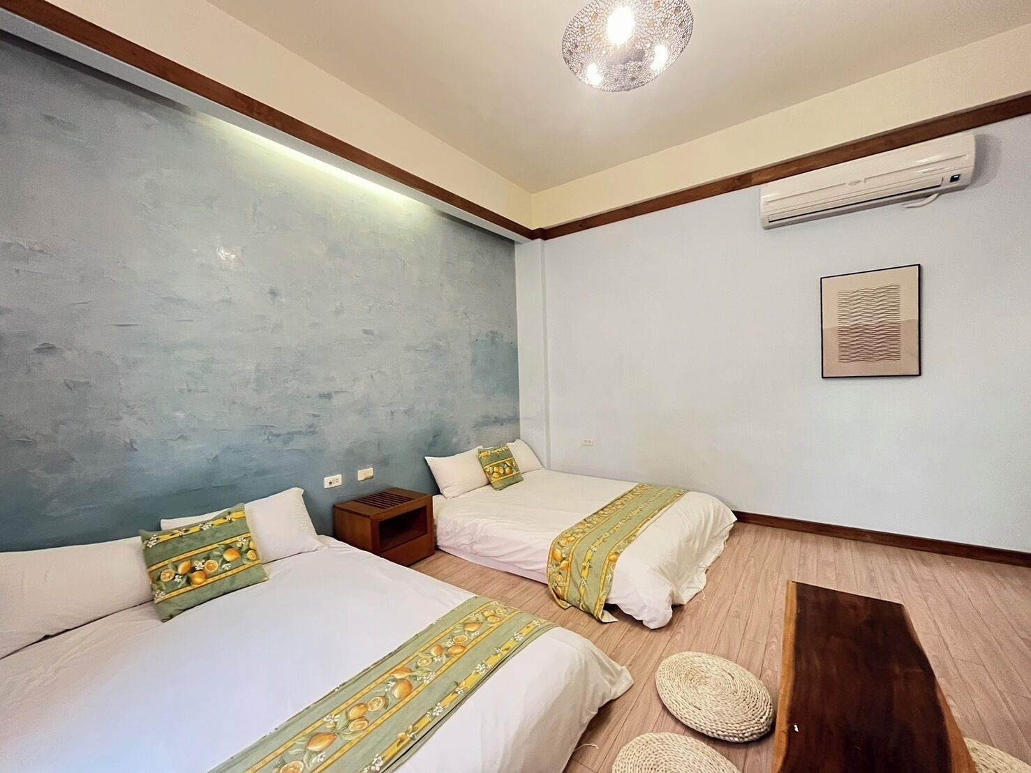 A room with two beds, a wooden floor, an air conditioning unit, a wall-mounted artwork, and a textured ceiling light.