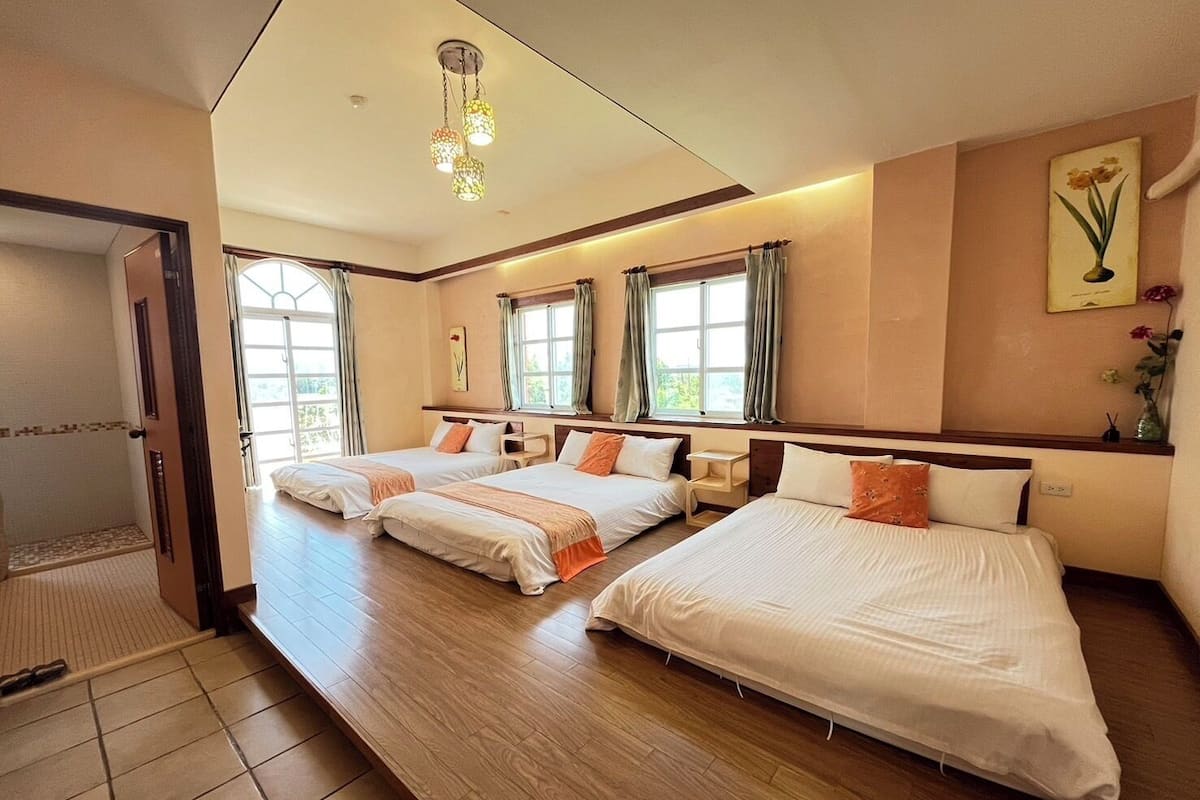 A hotel room with two beds, a wooden floor, a window with curtains, a bedside table, and a hanging light fixture.