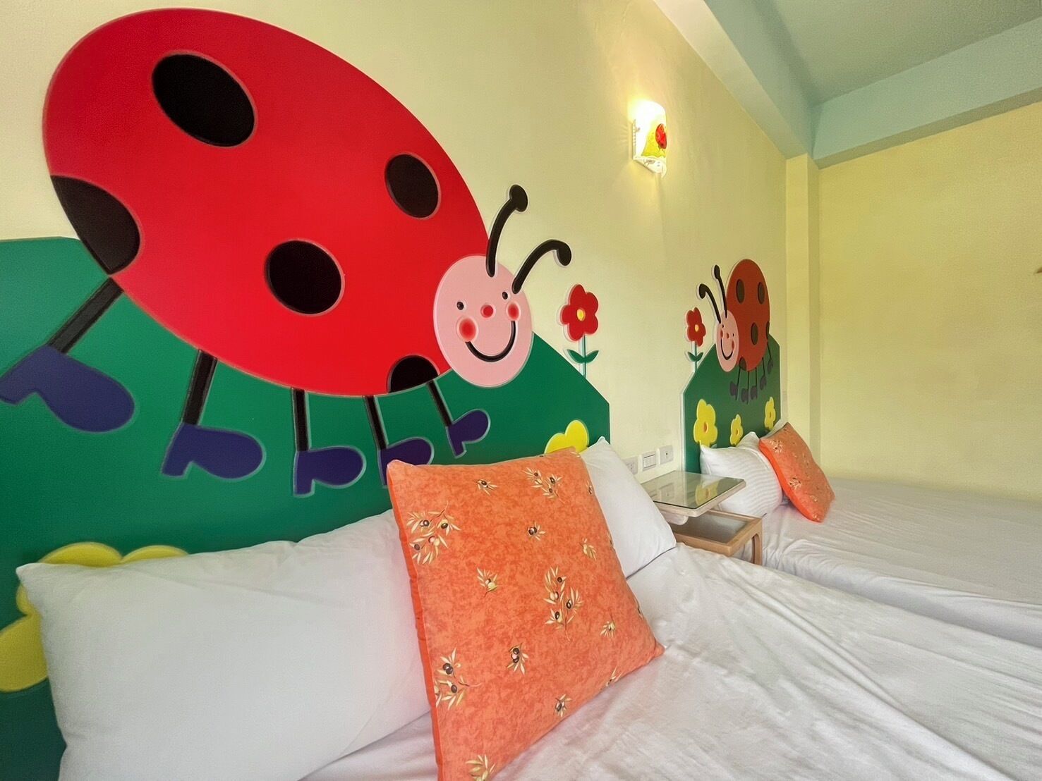Hotellihuone, jossa on sänky, tyynyjä, yöpöytä ja seinällä koristeellinen kuva leppäpirkosta.
