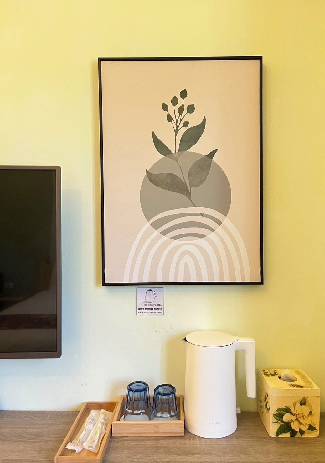A framed artwork of a plant on a wall, a white kettle, two glasses, and a tissue box on a wooden tray.