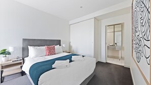 A modern hotel room with a large bed, white bedding, a grey headboard, and a blue throw blanket. There is a bedside table with a lamp, a small plant, and a book titled "Paris Style Guide." The room has a grey carpet and a white door leading to a bathroom with a mirror and a sink.