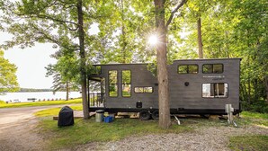 A mobile home with multiple windows and a covered porch, situated in a wooded area near a lake. - Tiny RV Escape with Stunning Lake Views, AC, WiFi (Nevis)