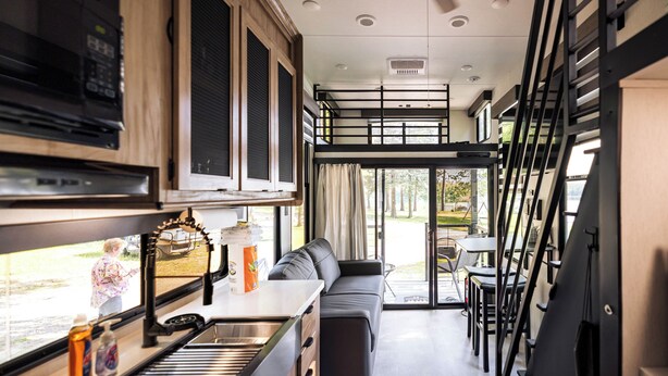 A compact kitchen with a microwave, stove, and sink. There is a sofa and a dining table with chairs. A window provides a view of the outdoors. - Tiny RV Escape with Stunning Lake Views, AC, WiFi (Nevis)