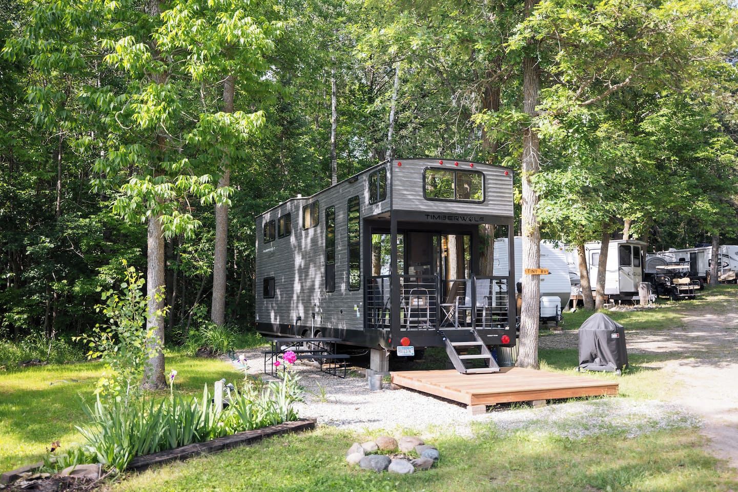 A mobile home with a deck, surrounded by trees and other RVs.