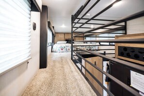 Un pasillo estrecho con una cama al final, una ventana con persianas y una pequeña cocina con armarios y microondas.