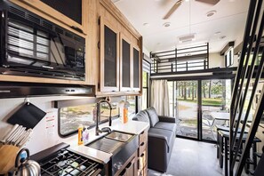 A compact kitchen with a stove, sink, and cabinets, a dining area with a table and chairs, and a view of the outdoors through large windows.