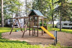 A playground with a swing set and a slide, surrounded by trees and RVs.